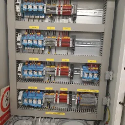 Otwarta szara szafa sterownicza z widocznymi bezpiecznikami, przekaźnikami, listwami zaciskowymi i okablowaniem, oznaczona żółtymi etykietami, z przewodami w dolnej części szafy.