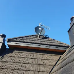 Antena satelitarna Invertio i antena naziemna zamontowane na dachu krytym brązową dachówką bitumiczną, widok z dołu na tle błękitnego nieba.