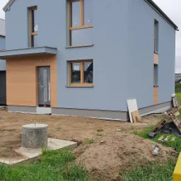 Dwukondygnacyjny dom z szarą elewacją i drewnianymi ramami okiennymi, z drewnianą okładziną wejściową, na etapie wykończenia, widoczne materiały budowlane i wykop ziemny.