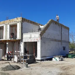 Budowa domu jednorodzinnego w stanie surowym otwartym: widoczne ściany z bloczków, szalunki na stropie, pracownicy na rusztowaniu i dachu, oraz materiały budowlane na placu budowy w słoneczny dzień.