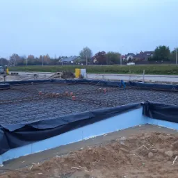 Przygotowanie zbrojenia fundamentów na placu budowy: pracownik w odblaskowej kamizelce łączy druty zbrojeniowe na siatce, widoczne niebieskie płyty izolacyjne i wystające pomarańczowe rury, w tle...