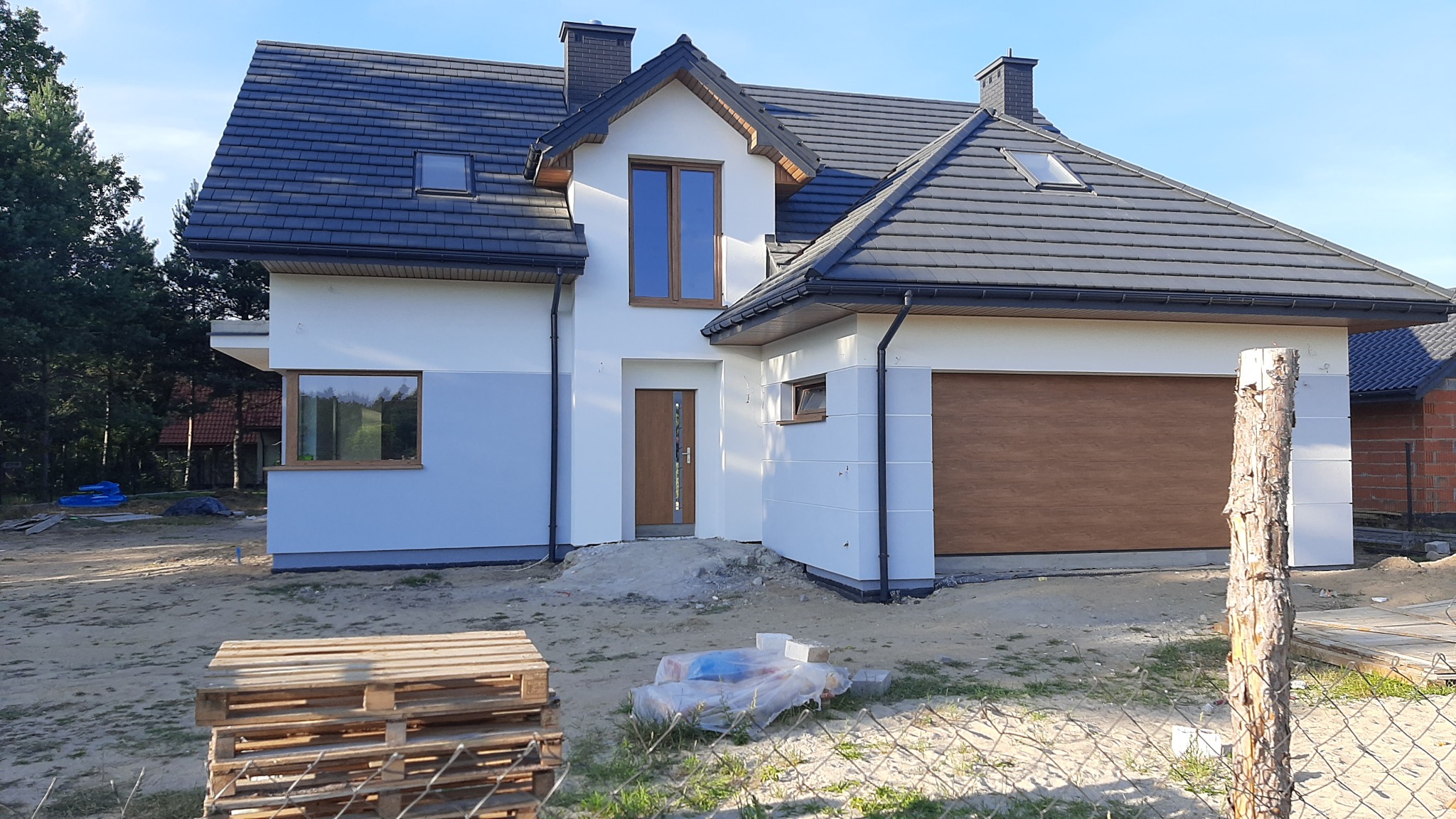 Nowoczesny dom jednorodzinny z szarą dachówką i białą elewacją, częściowo pomalowaną na szaro, z drewnianymi oknami i bramą garażową, widoczny z zewnątrz, w trakcie prac wykończeniowych.