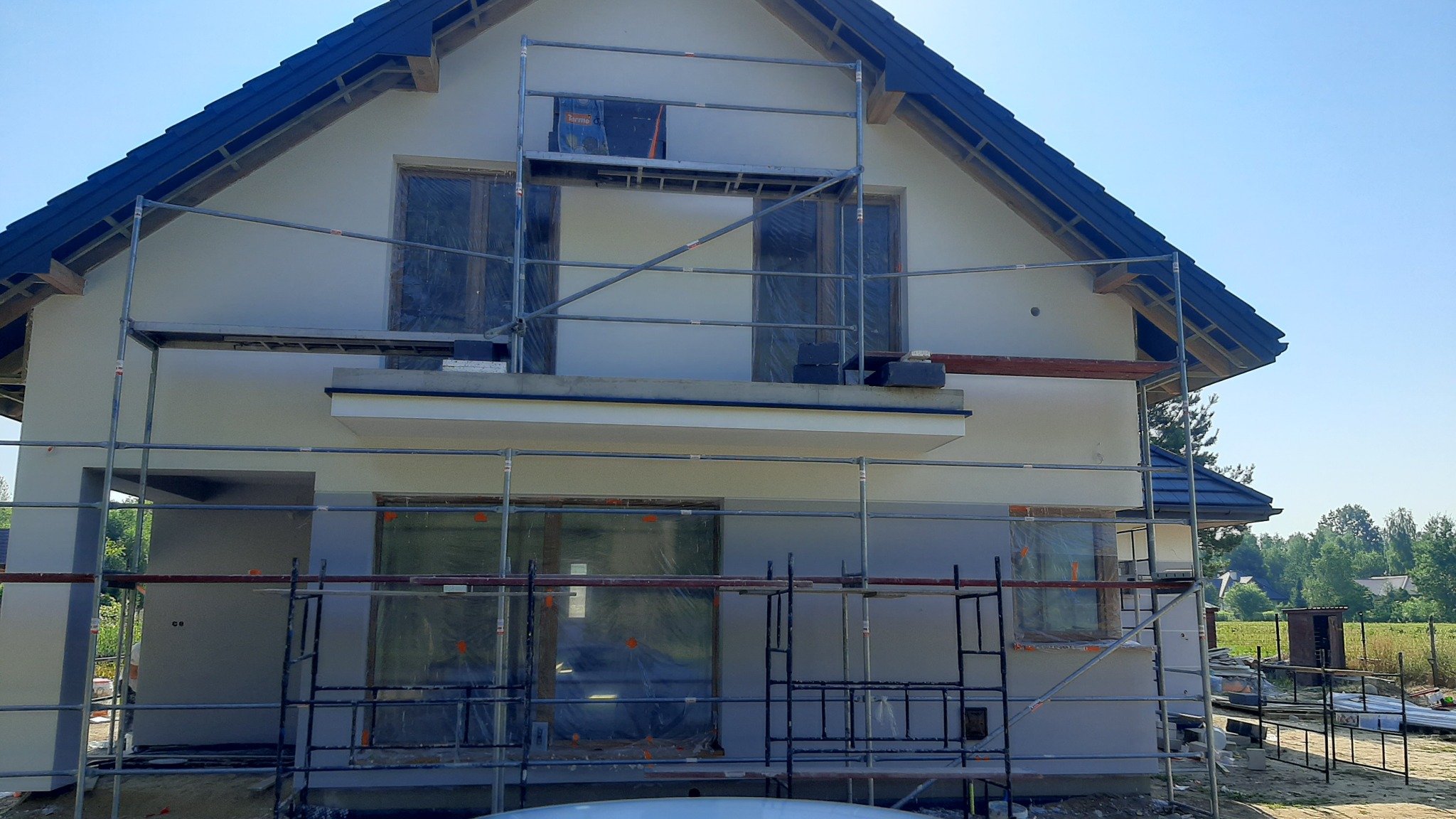 Dom jednorodzinny w trakcie prac elewacyjnych, okna zabezpieczone folią, widoczne rusztowania, świeżo nałożony tynk w jasnym odcieniu szarości, niebieska dachówka, błękitne niebo