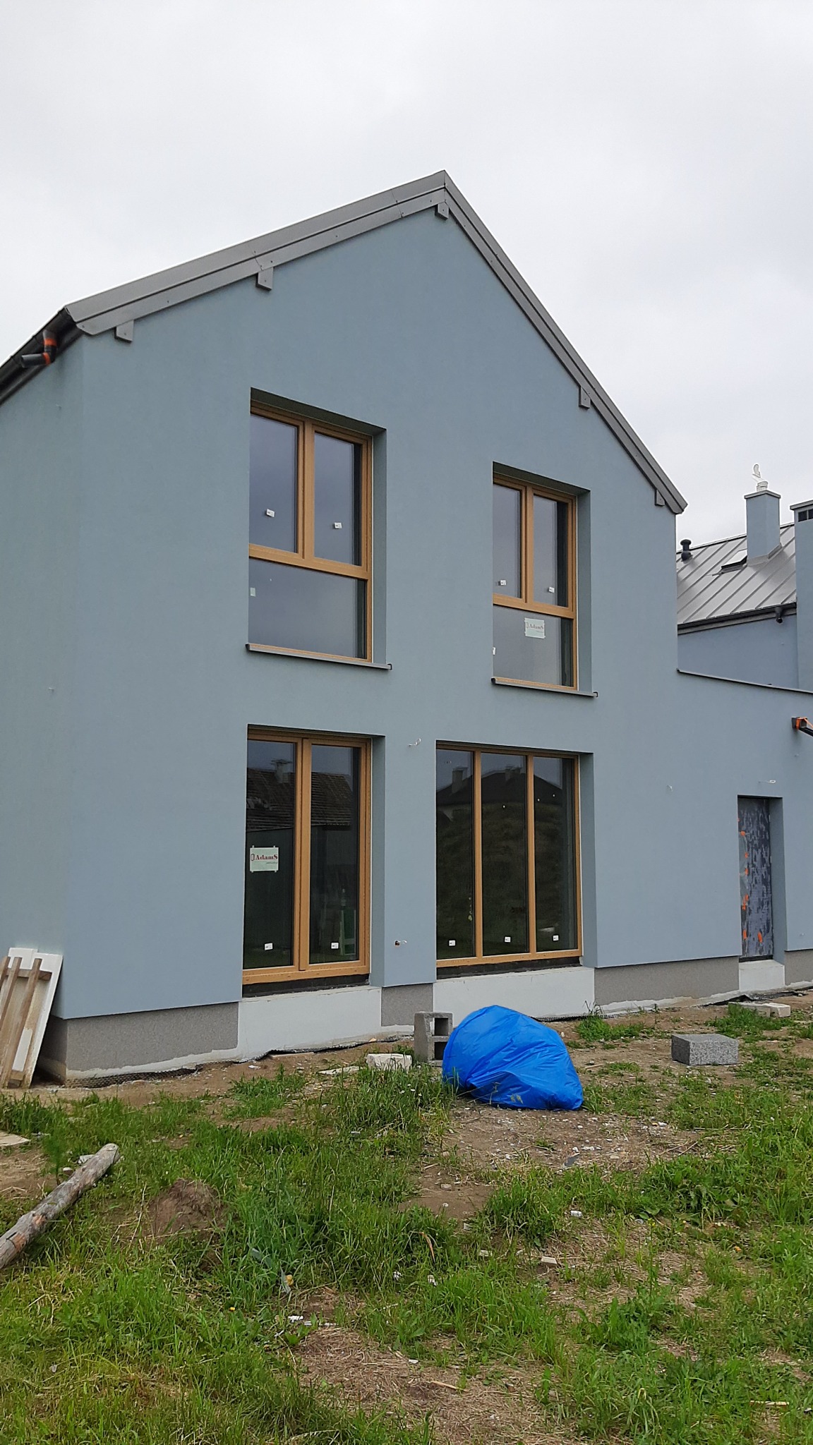 Dwukondygnacyjny dom z szarą elewacją i drewnianymi oknami, widoczne niedokończone prace wykończeniowe wokół budynku, w tym niebieski worek z materiałami budowlanymi.