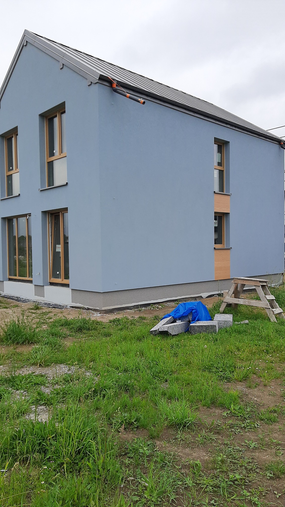 Nowoczesny, dwukondygnacyjny dom z szarą elewacją, drewnianymi ramami okiennymi i metalowym dachem. Na trawniku leżą bloczki budowlane przykryte niebieską folią i drewniany kozioł.