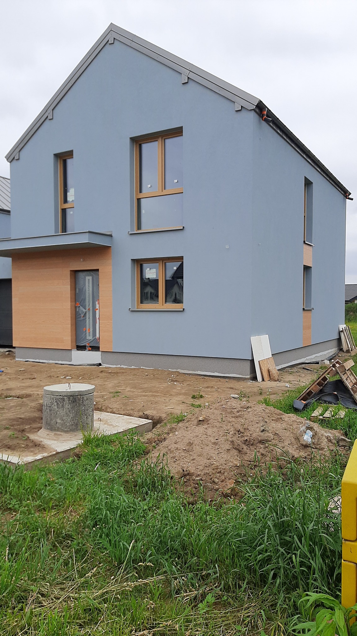 Dwukondygnacyjny dom z szarą elewacją i drewnianymi ramami okiennymi, z drewnianą okładziną wejściową, na etapie wykończenia, widoczne materiały budowlane i wykop ziemny.