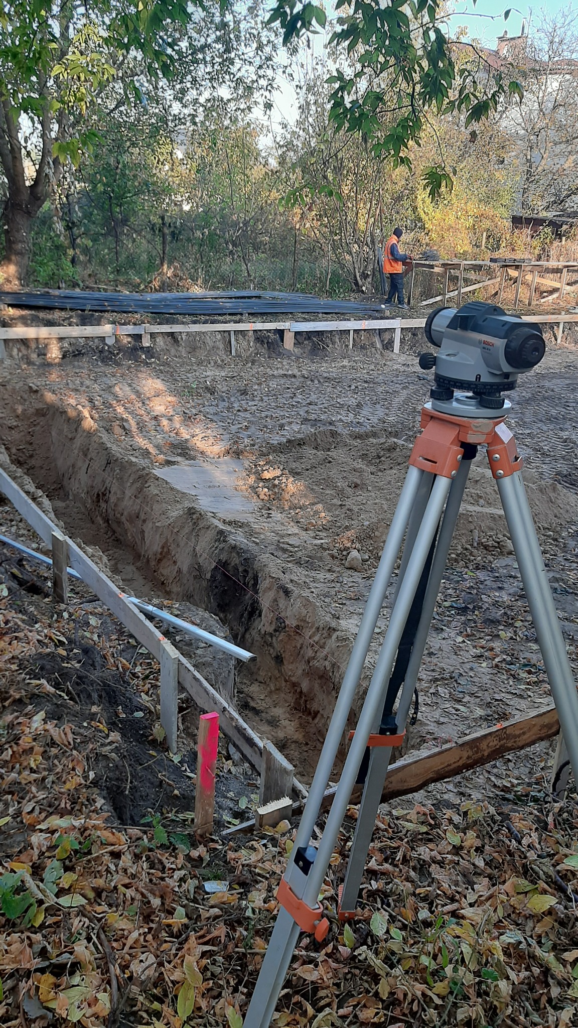 Wykop pod fundamenty z szalunkiem, widoczny niwelator optyczny na statywie, w tle pracownik w kamizelce odblaskowej.