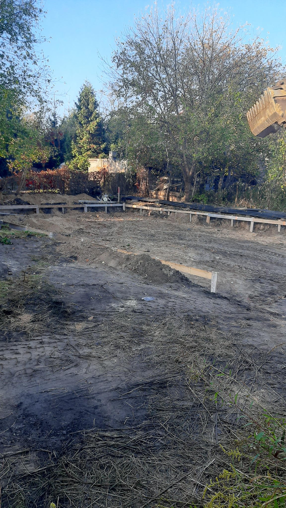 Przygotowany teren pod fundamenty z widocznym szalunkiem i częściowo wyrównaną ziemią, w tle drzewa i fragment koparki.