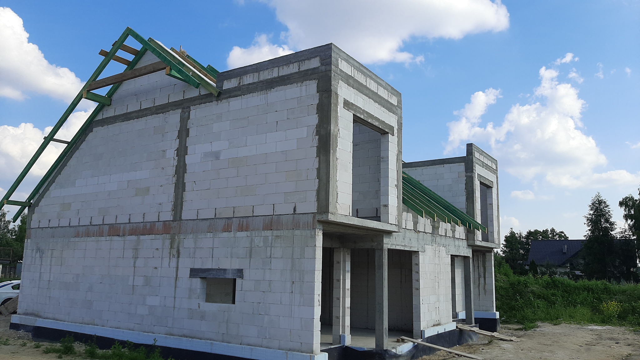 Surowy stan dwukondygnacyjnego domu z białych bloczków, z widoczną drewnianą konstrukcją dachu pomalowaną na zielono, na tle błękitnego nieba z chmurami.