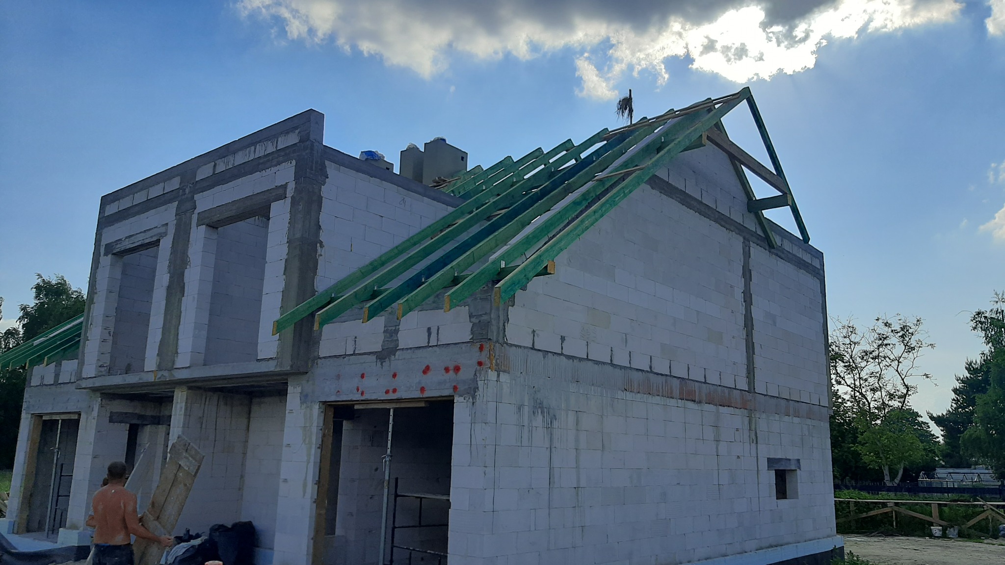 Niedokończony dwukondygnacyjny dom z białego bloczka, z zielonymi krokwiami dachowymi, widok z zewnątrz, w tle błękitne niebo z chmurami.