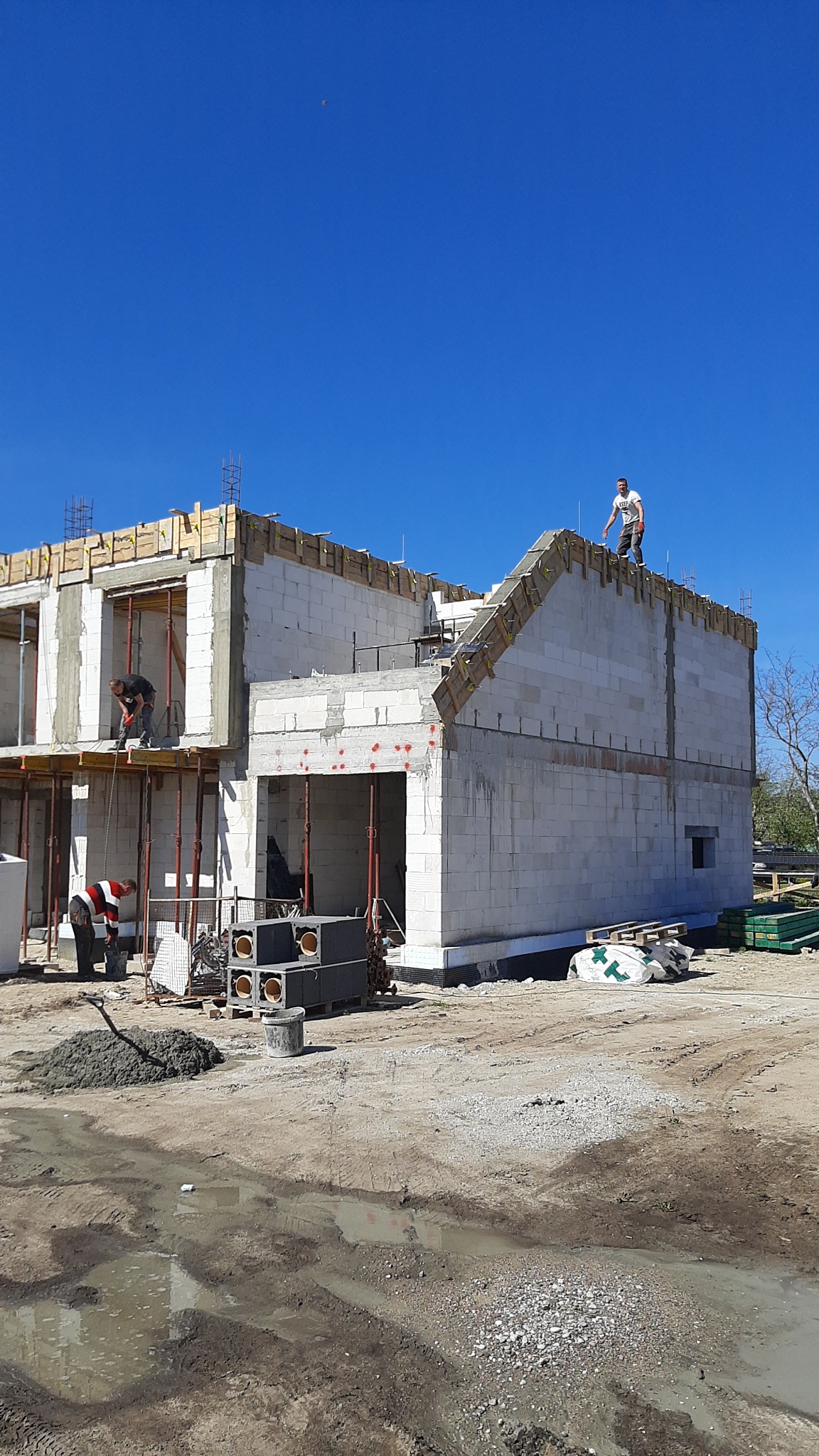 Budowa domu jednorodzinnego w stanie surowym otwartym: widoczne ściany z bloczków, szalunki na stropie, pracownicy na rusztowaniu i dachu, oraz materiały budowlane na placu budowy w słoneczny dzień.