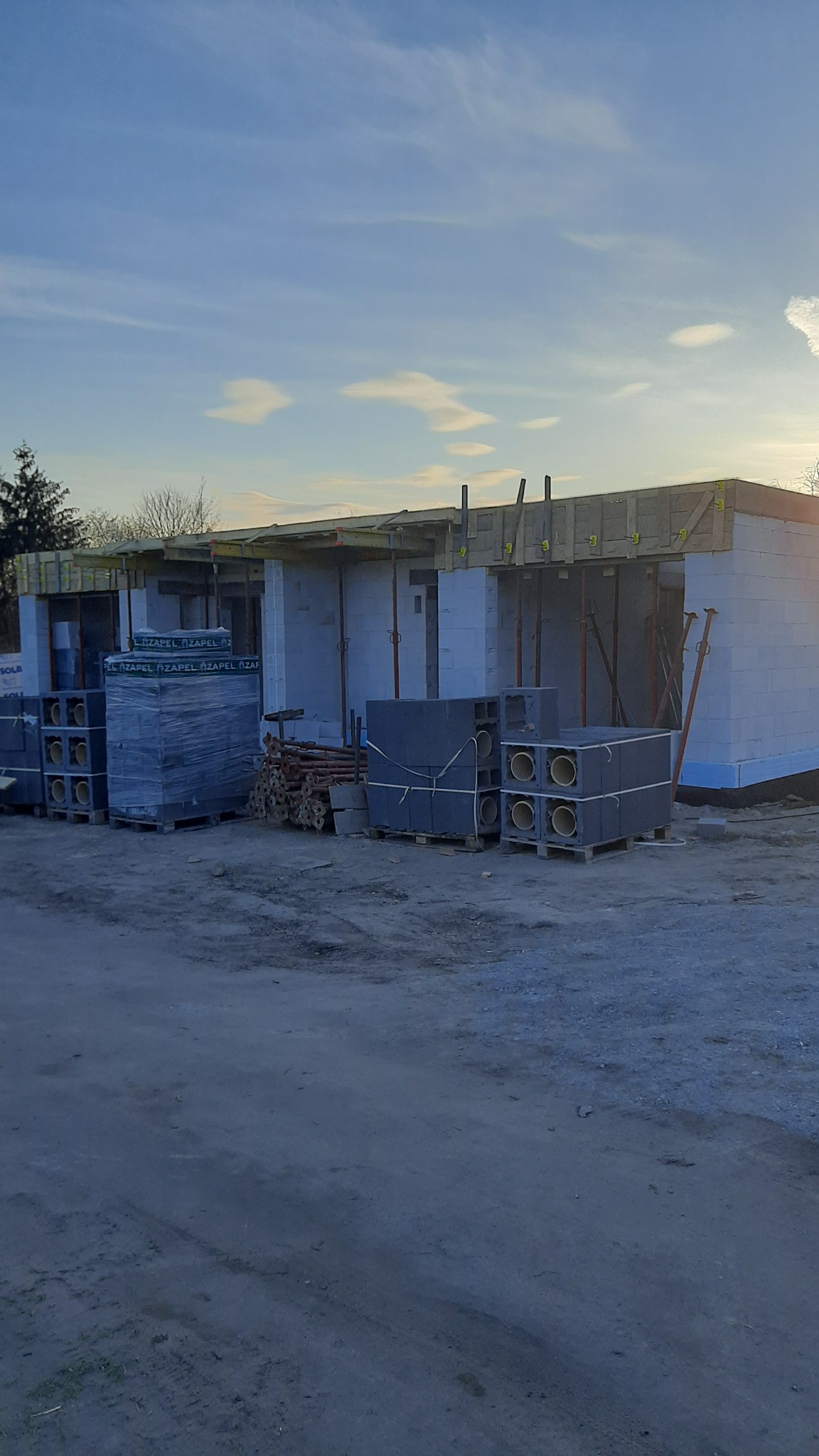 Budowa domu jednorodzinnego w trakcie wznoszenia ścian z bloczków, widoczne materiały budowlane, podpory stropowe i niebo w tle.