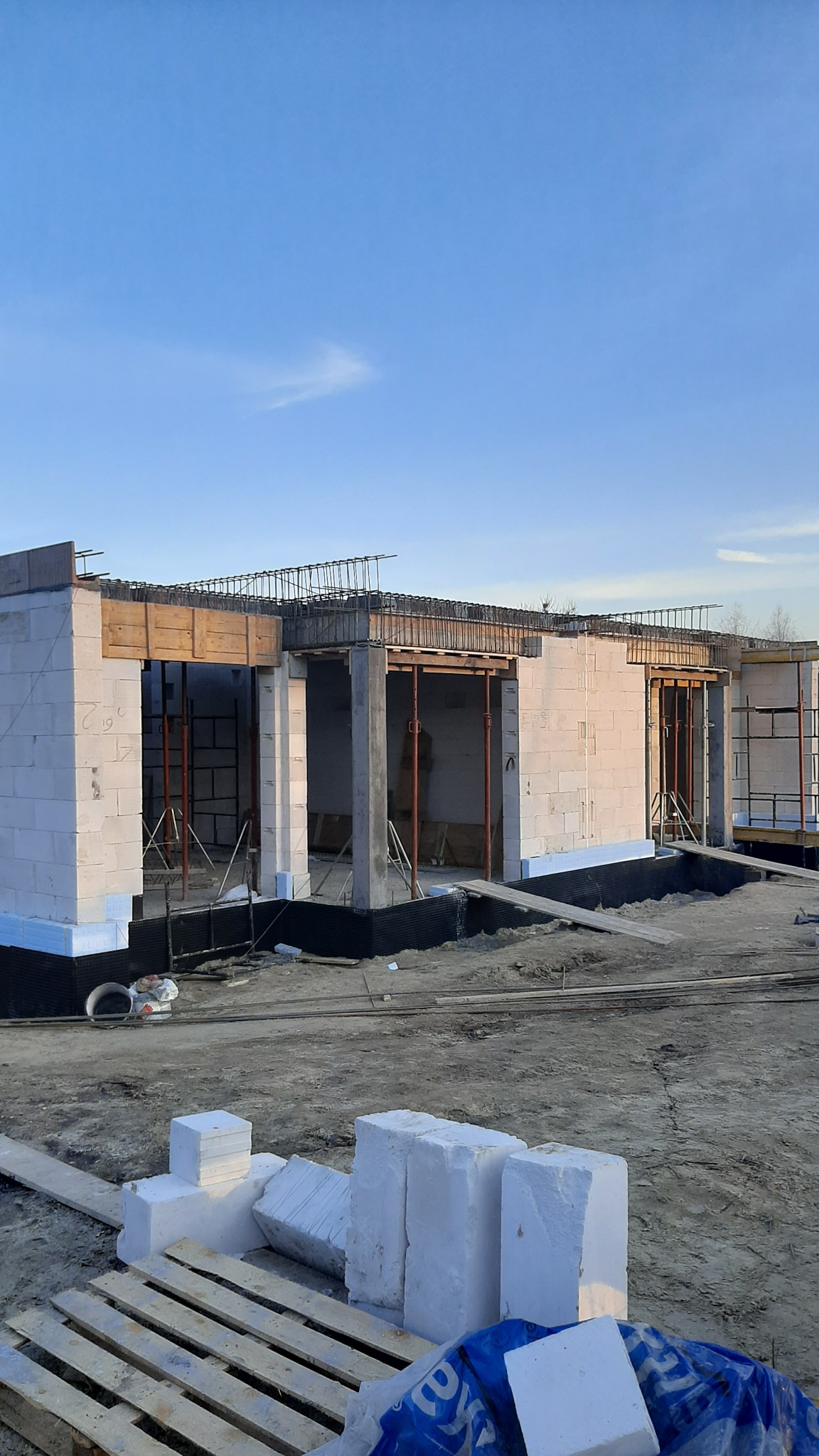 Budowa domu jednorodzinnego: wznoszenie ścian z białych bloczków, widoczne szalunki i zbrojenia, fundament ocieplony styropianem, teren budowy w trakcie prac.