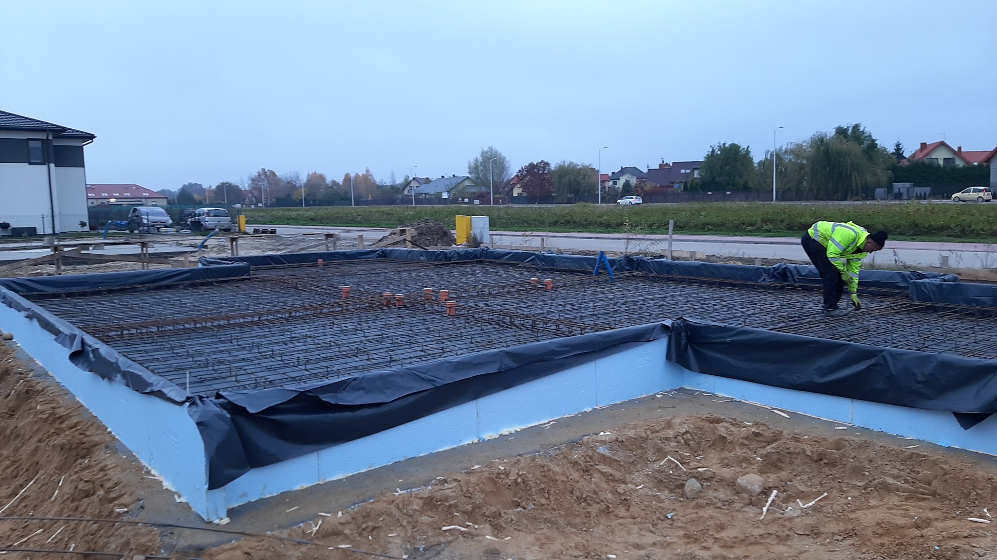 Przygotowanie zbrojenia fundamentów na placu budowy: pracownik w odblaskowej kamizelce łączy druty zbrojeniowe na siatce, widoczne niebieskie płyty izolacyjne i wystające pomarańczowe rury, w tle...