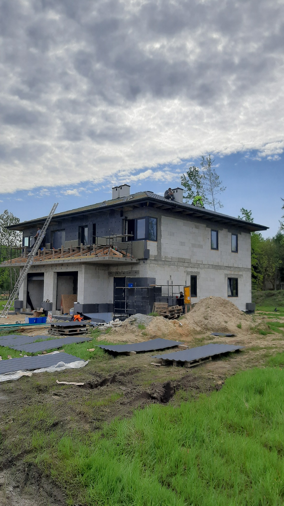 Dwupiętrowy dom w trakcie budowy z szarego bloczka, widoczny montaż elewacji, rusztowania, drabina oparta o ścianę, robotnicy na dachu i przy wejściu, stosy materiałów budowlanych na ziemi.