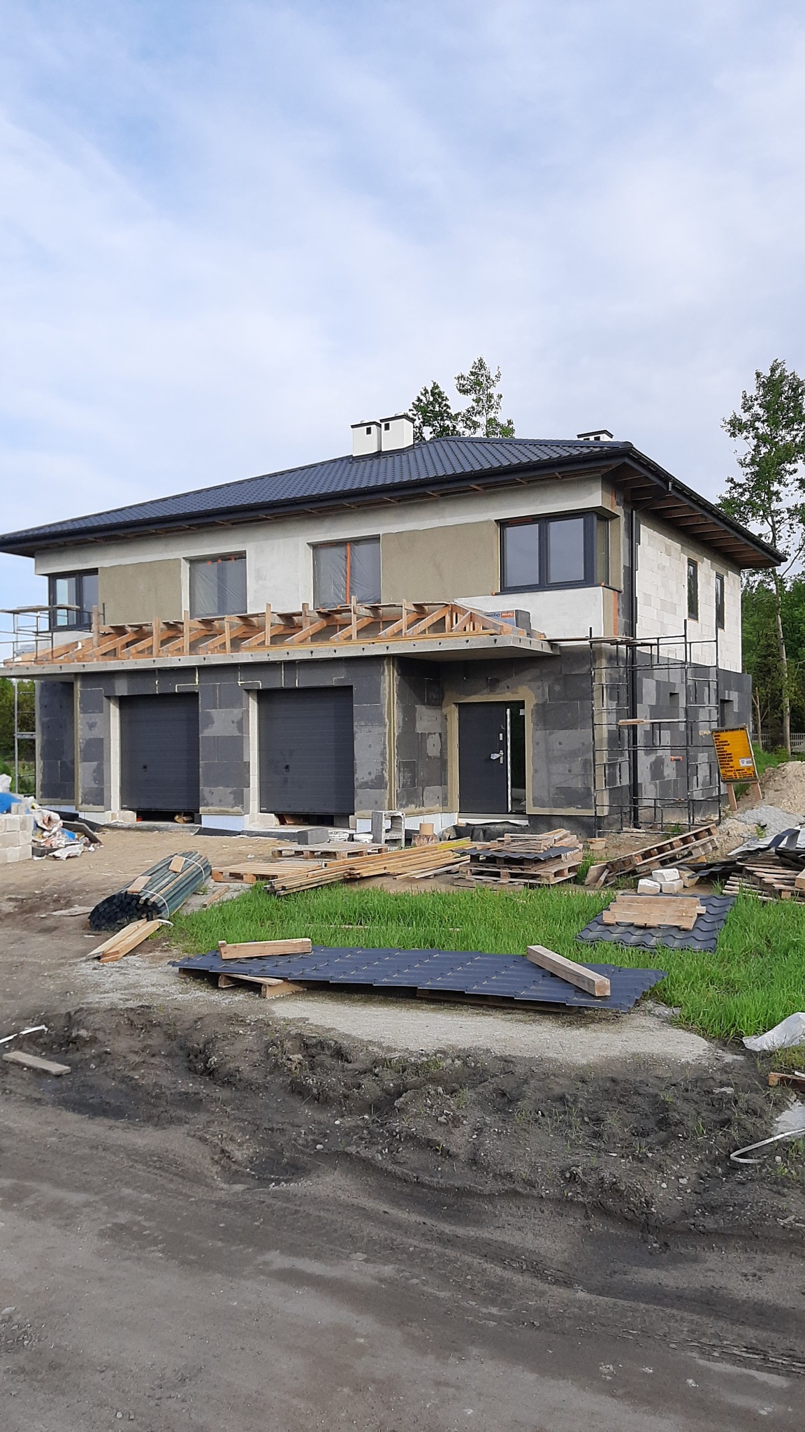 Dwukondygnacyjny dom w trakcie budowy z widocznymi elementami konstrukcyjnymi, szarą elewacją w dolnej części, częściowo otynkowaną górną kondygnacją i drewnianą konstrukcją nad garażem. Na ziemi...