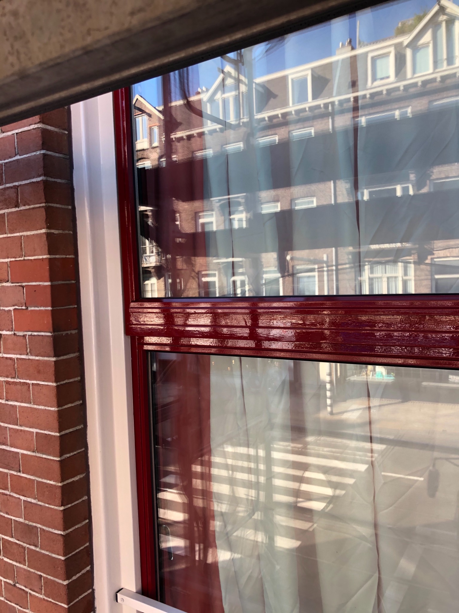Fragment okna z pomalowaną na bordowo ramą i parapetem, widoczna ceglana ściana budynku oraz odbicie sąsiedniego budynku w szybie.