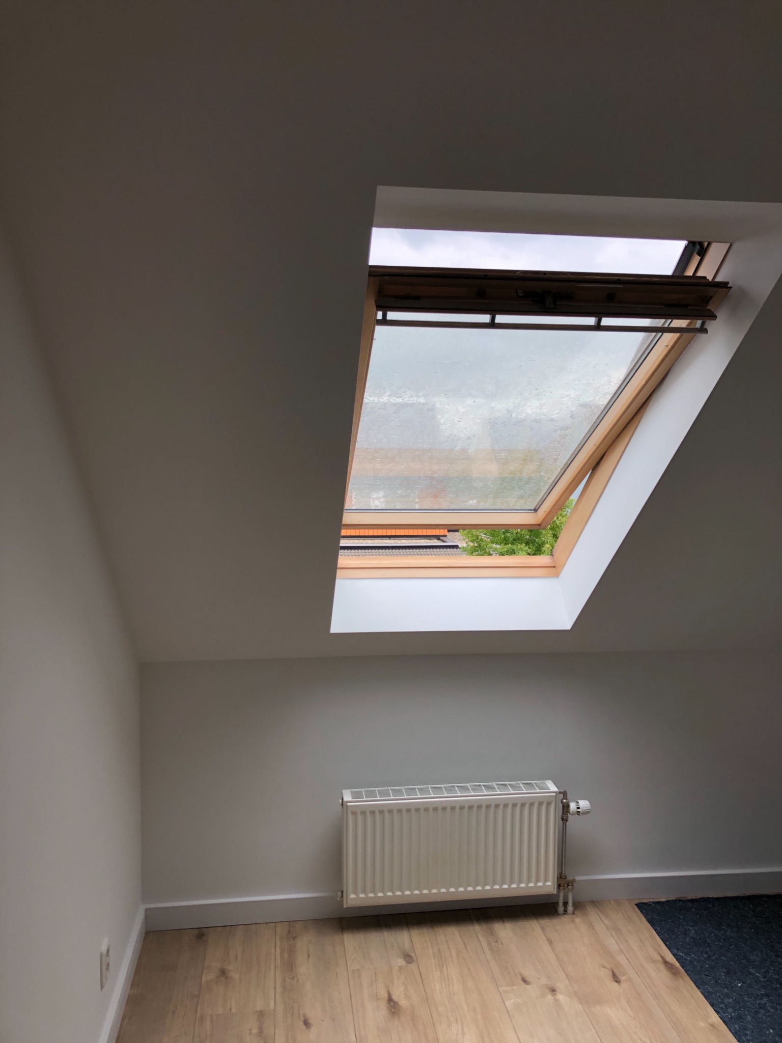 Poddasze po malowaniu z oknem dachowym uchylonym do góry, grzejnik pomalowany na biało, parkiet w odcieniach beżu
