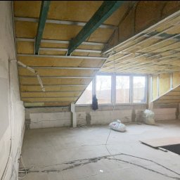 Remo-pats - Poddasze w trakcie adaptacji: widoczne ocieplenie wełną mineralną w konstrukcji sufitu, ściany z bloczków, instalacje elektryczne na podłodze, okna dachowe z widokiem na zabudowę jednorodzinną.