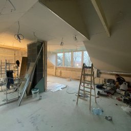 Remo-pats - Poddasze w trakcie remontu z widoczną konstrukcją sufitu z płyt kartonowo-gipsowych, rusztowaniem, rozłożonymi narzędziami, oknami i pracującym mężczyzną w oddali.