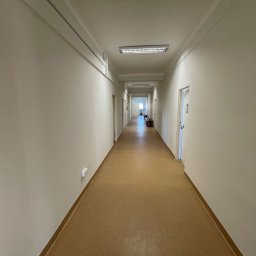 Remo-pats - Długi, jasny korytarz z beżowymi ścianami i podłogą z wykładziny. Widoczne drzwi po obu stronach i świetlówki na suficie. Perspektywa zbieżna, na końcu korytarza światło dzienne.