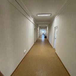 Remo-pats - Długi korytarz z beżowymi ścianami i brązową podłogą. Białe drzwi po obu stronach. Oświetlenie sufitowe wzdłuż korytarza. Perspektywa zbieżna w oddali.