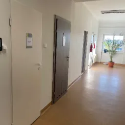 Korytarz biurowy z dwiema parami drzwi, pomalowanymi na biało ścianami i brązową podłogą. W oddali widoczne okno z rośliną doniczkową. Oświetlenie sufitowe.