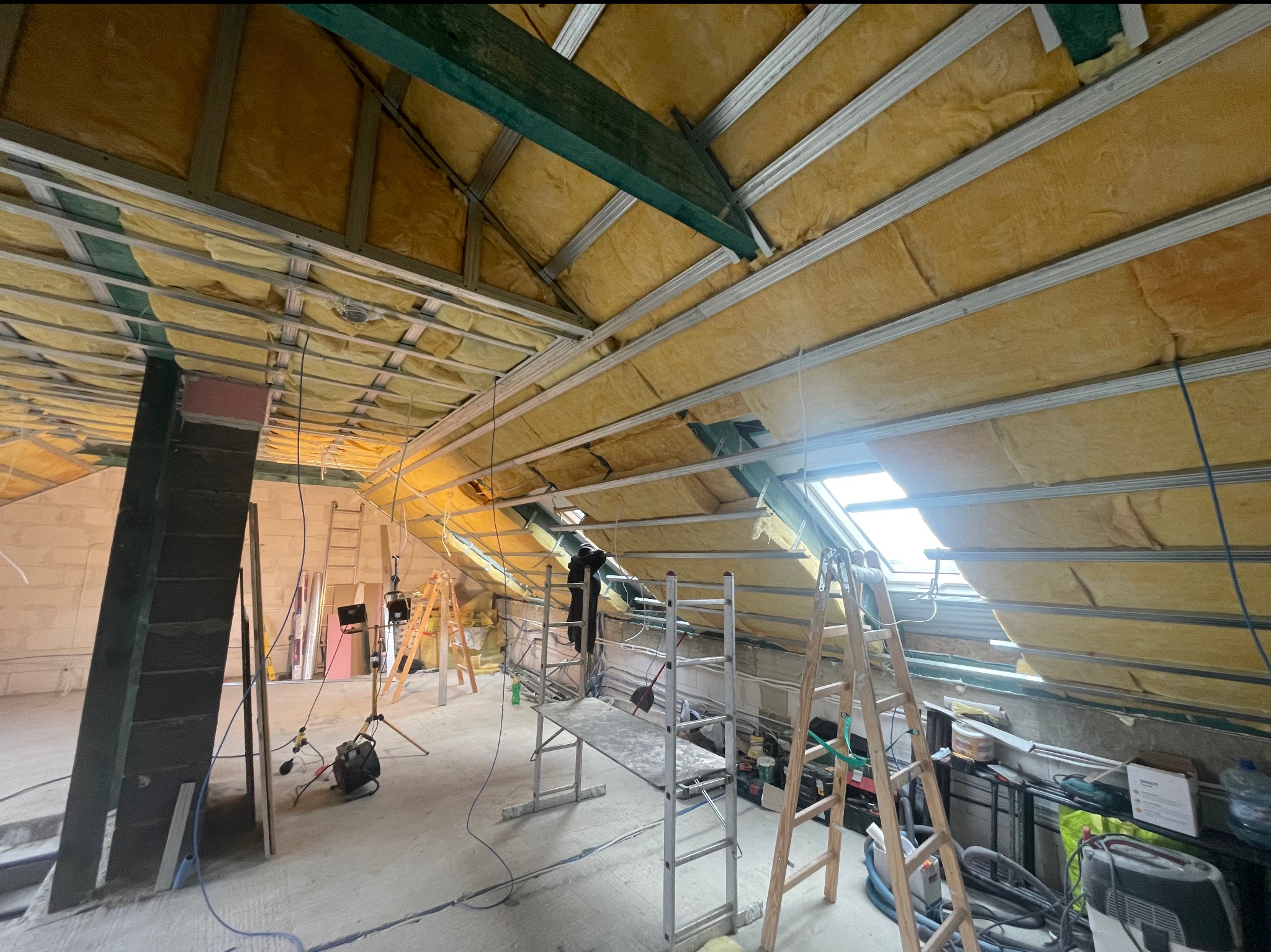 Montaż izolacji z wełny mineralnej na poddaszu, widoczne metalowe profile, rusztowanie i drabiny, okno dachowe w tle.