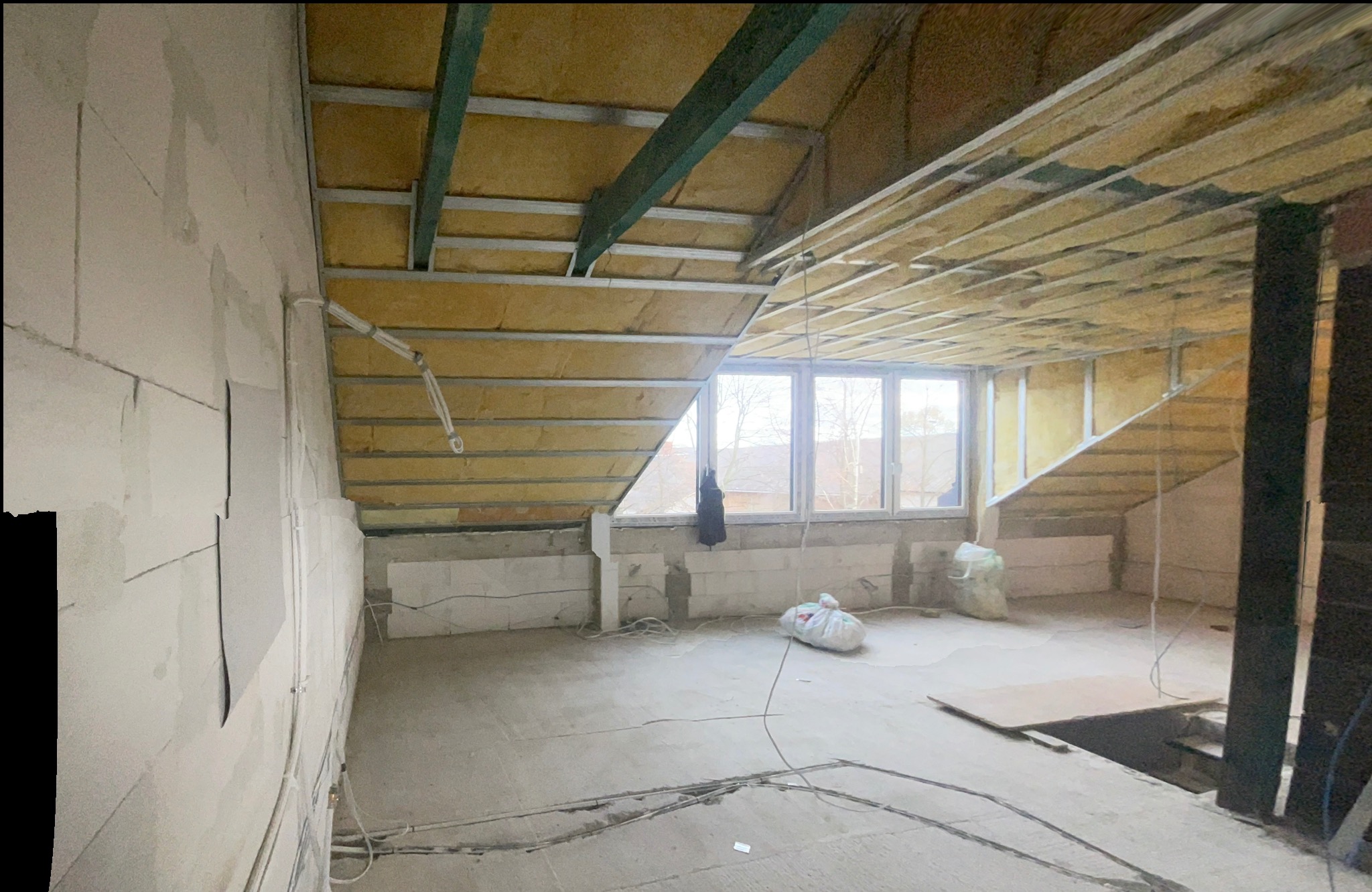 Poddasze w trakcie adaptacji: widoczne ocieplenie wełną mineralną w konstrukcji sufitu, ściany z bloczków, instalacje elektryczne na podłodze, okna dachowe z widokiem na zabudowę jednorodzinną.