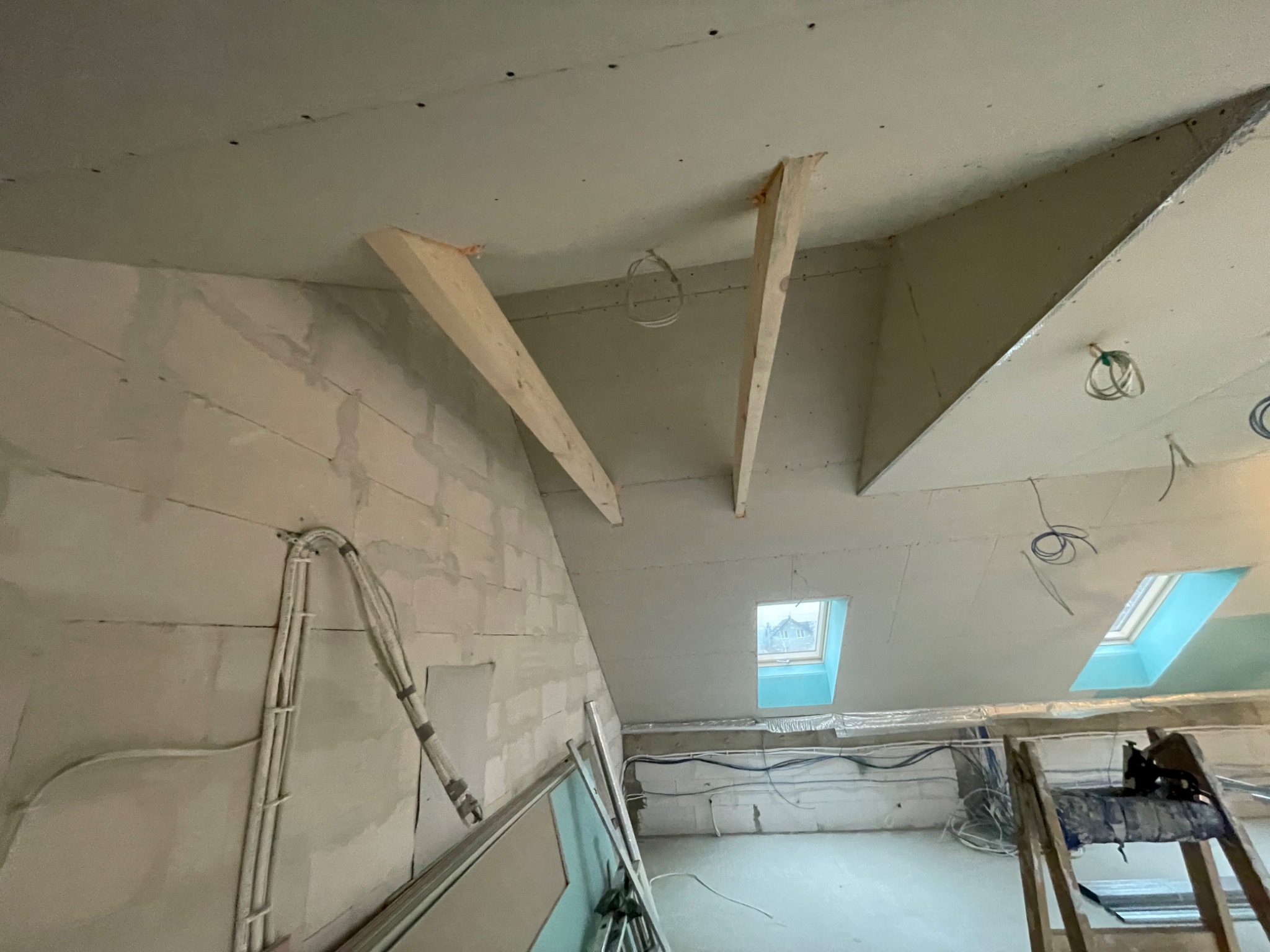 Poddasze w trakcie remontu: widoczne drewniane wzmocnienia sufitu, ściany z surowego betonu komórkowego, przewody elektryczne i okna dachowe, fragment drabiny malarskiej.