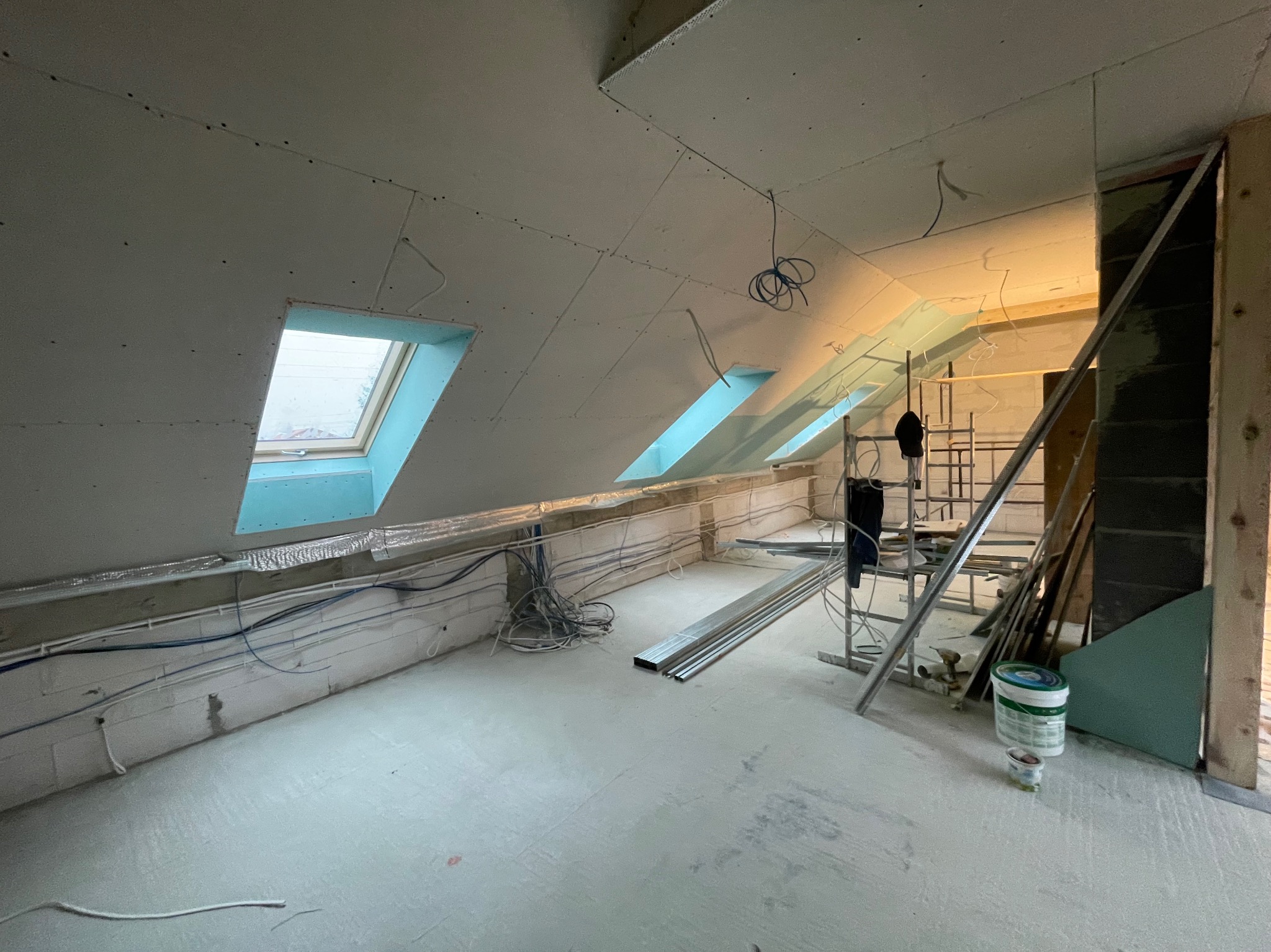 Poddasze w trakcie remontu: zamontowane płyty kartonowo-gipsowe na skosach dachu z oknami, widoczne przewody elektryczne, aluminiowe profile, rusztowanie i wiadro z farbą.