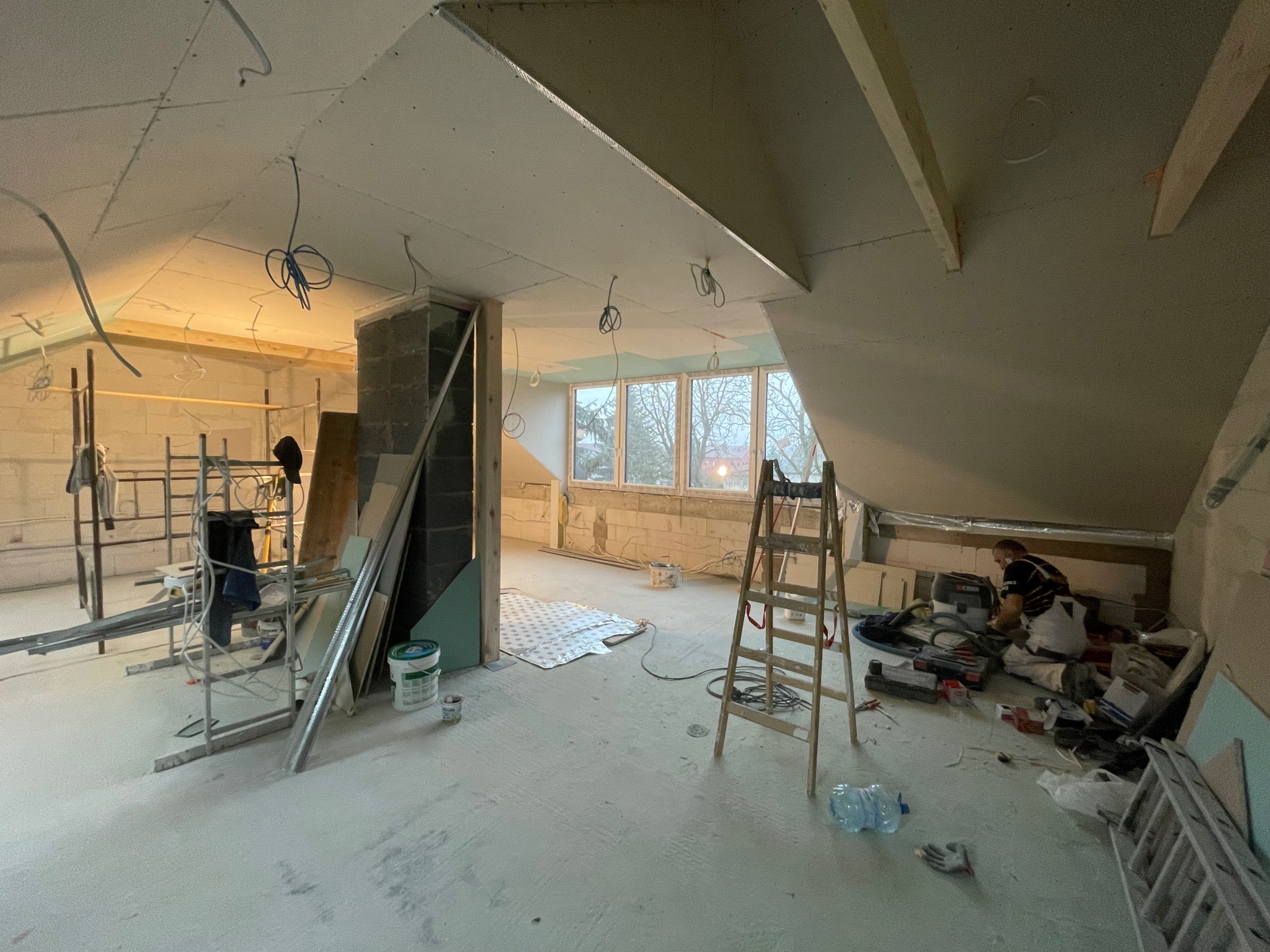 Poddasze w trakcie remontu z widoczną konstrukcją sufitu z płyt kartonowo-gipsowych, rusztowaniem, rozłożonymi narzędziami, oknami i pracującym mężczyzną w oddali.