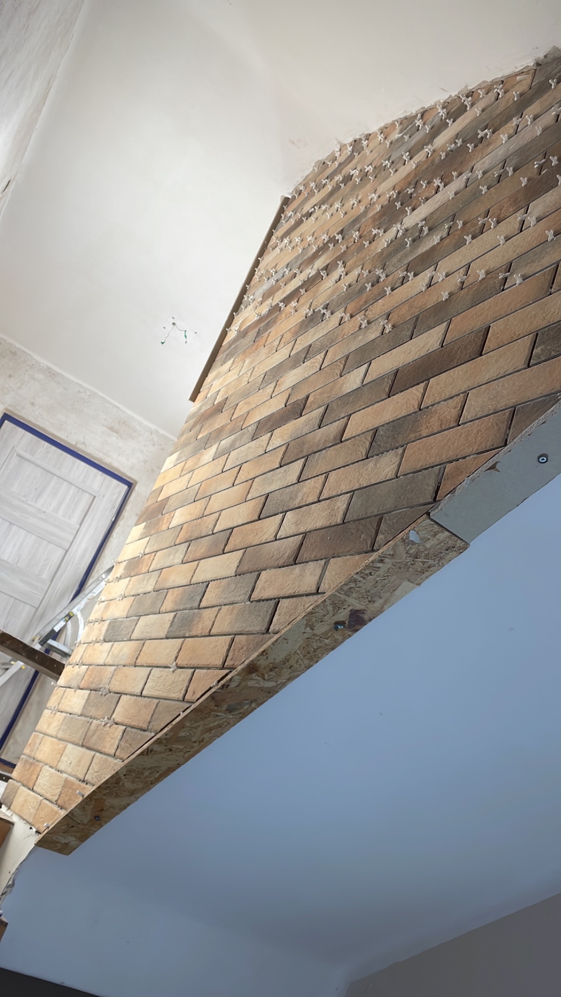 Ściana wykończona brązowymi, prostokątnymi płytkami z krzyżykami dystansowymi, widoczne elementy konstrukcyjne i drabina. Układ ukośny płytek.