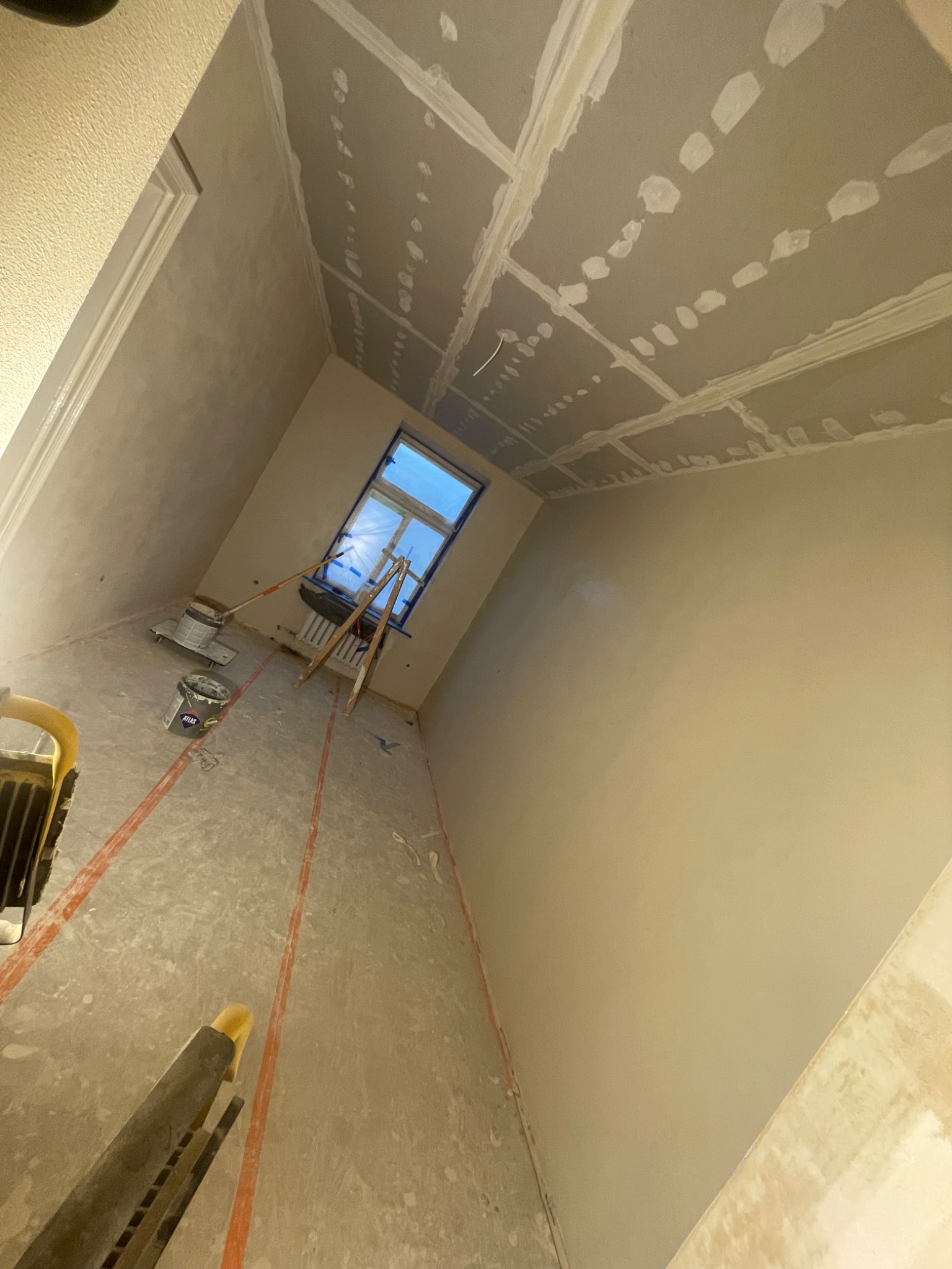 Pokój w trakcie prac wykończeniowych: ściany po szpachlowaniu, sufit z płyt kartonowo-gipsowych ze śladami szpachlowania. Widoczne narzędzia i drabina przy oknie.