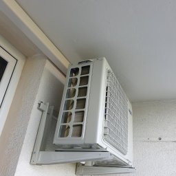 DECOMF Sp. z o. o. - Biała jednostka zewnętrzna klimatyzatora marki AUX zamontowana na ścianie budynku z widoczną fakturą tynku i fragmentem okna.