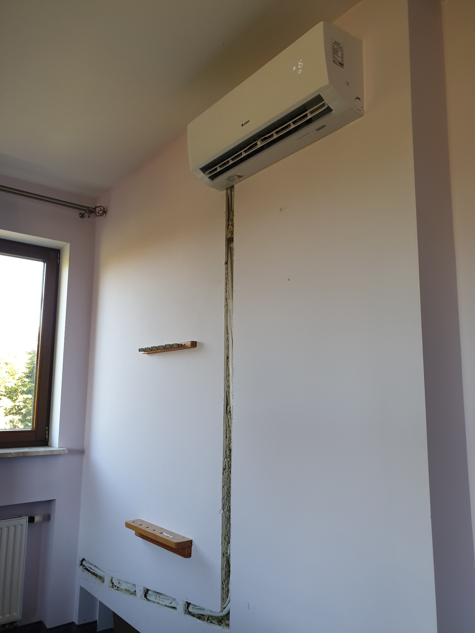 Zamontowany klimatyzator Gree na ścianie w trakcie instalacji, widoczne kable i otwory w ścianie, drewniane podpórki, okno z widokiem na zieleń.