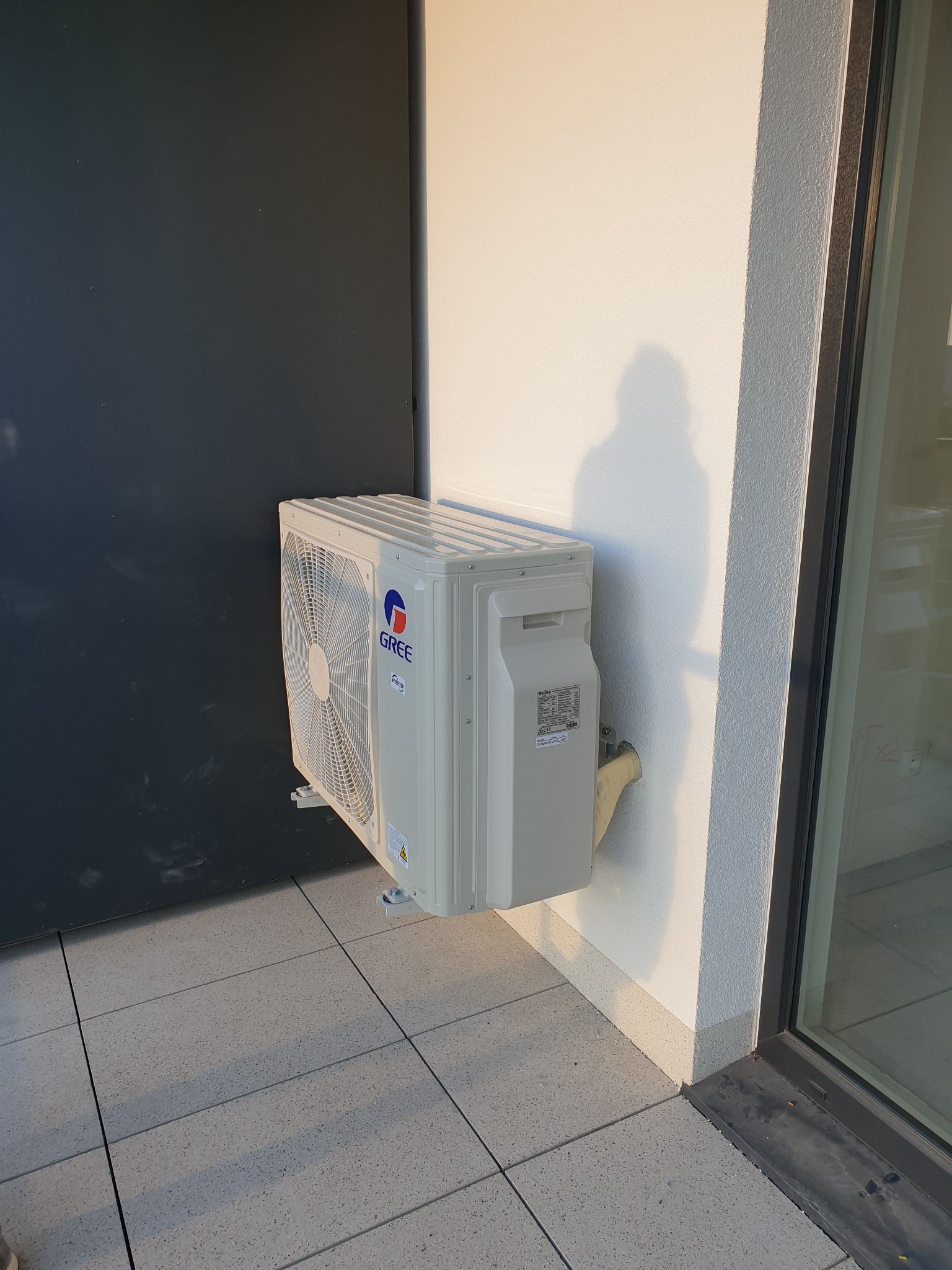 Zewnętrzna jednostka klimatyzacji Gree zamontowana na elewacji budynku, widoczne rury odprowadzające skropliny oraz cień osoby robiącej zdjęcie.