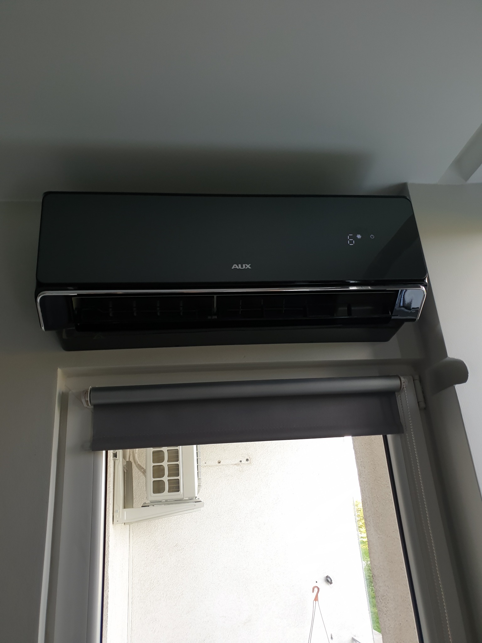 Czarny klimatyzator AUX zamontowany nad oknem z roletą, widoczny zewnętrzny moduł klimatyzacji na balkonie.
