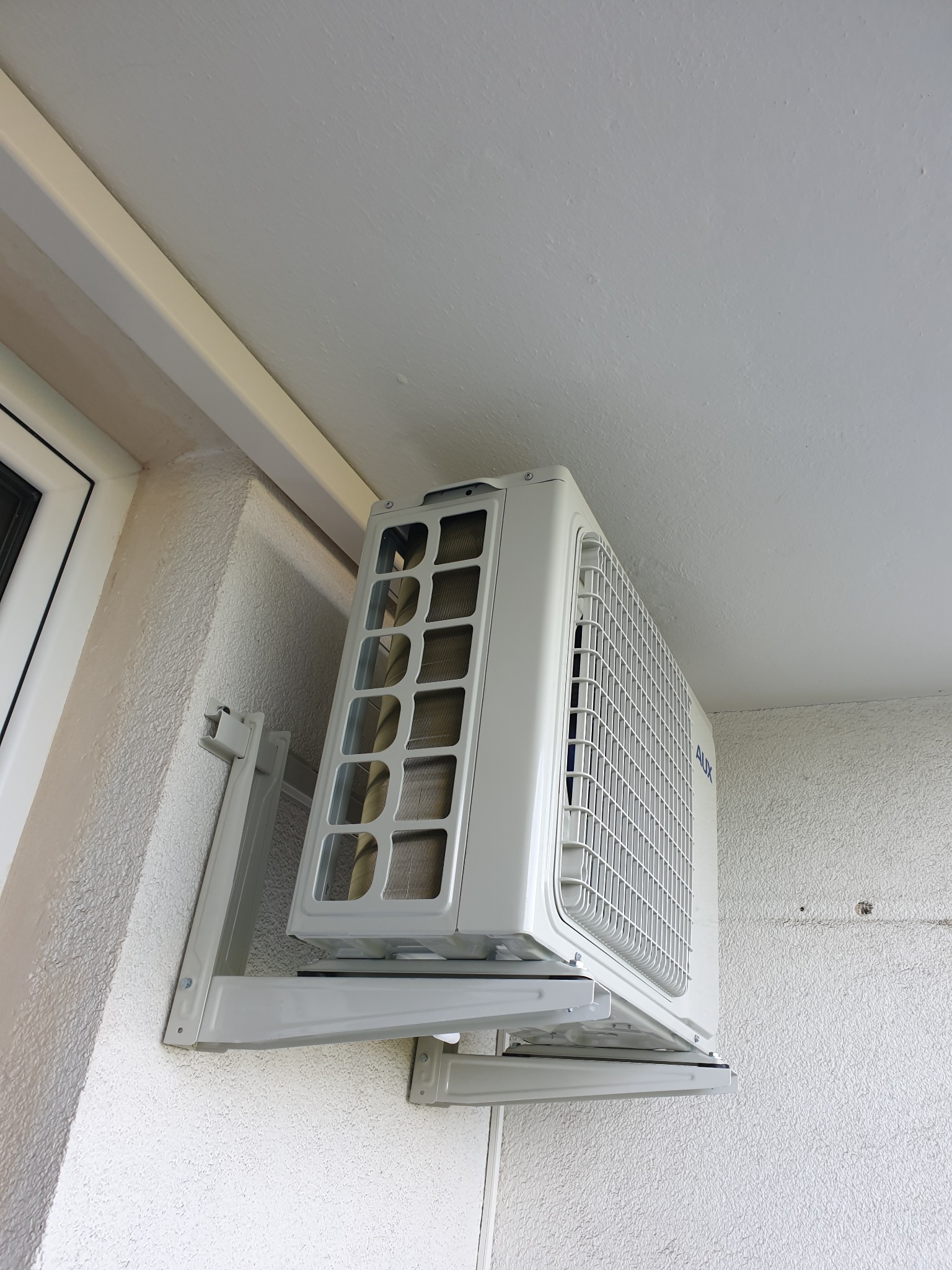 Biała jednostka zewnętrzna klimatyzatora marki AUX zamontowana na ścianie budynku z widoczną fakturą tynku i fragmentem okna.