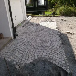 Fragment układanej ścieżki z kostki granitowej w kształcie łuku, widoczne przygotowane podłoże z grysu i zapas kostki, obok ściana budynku z rynną.