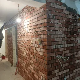 Montaż okładziny z cegły klinkierowej na ścianie wewnętrznej, widoczne krzyżyki dystansowe, świeżo położone płytki z szarą zaprawą, wystające kable elektryczne, poziomica oparta o ścianę.