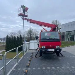 Czerwony podnośnik koszowy z pracownikiem w żółtej kamizelce, stojącym w koszu i pracującym przy słupie oświetleniowym na tle pochmurnego nieba i budynku.