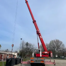 Czerwony dźwig samochodowy z wysuniętym ramieniem, przygotowany do podnoszenia czarnych pakunków na paletach na placu z widocznym boiskiem do koszykówki w tle, zabezpieczony barierkami ostrzegawczymi.