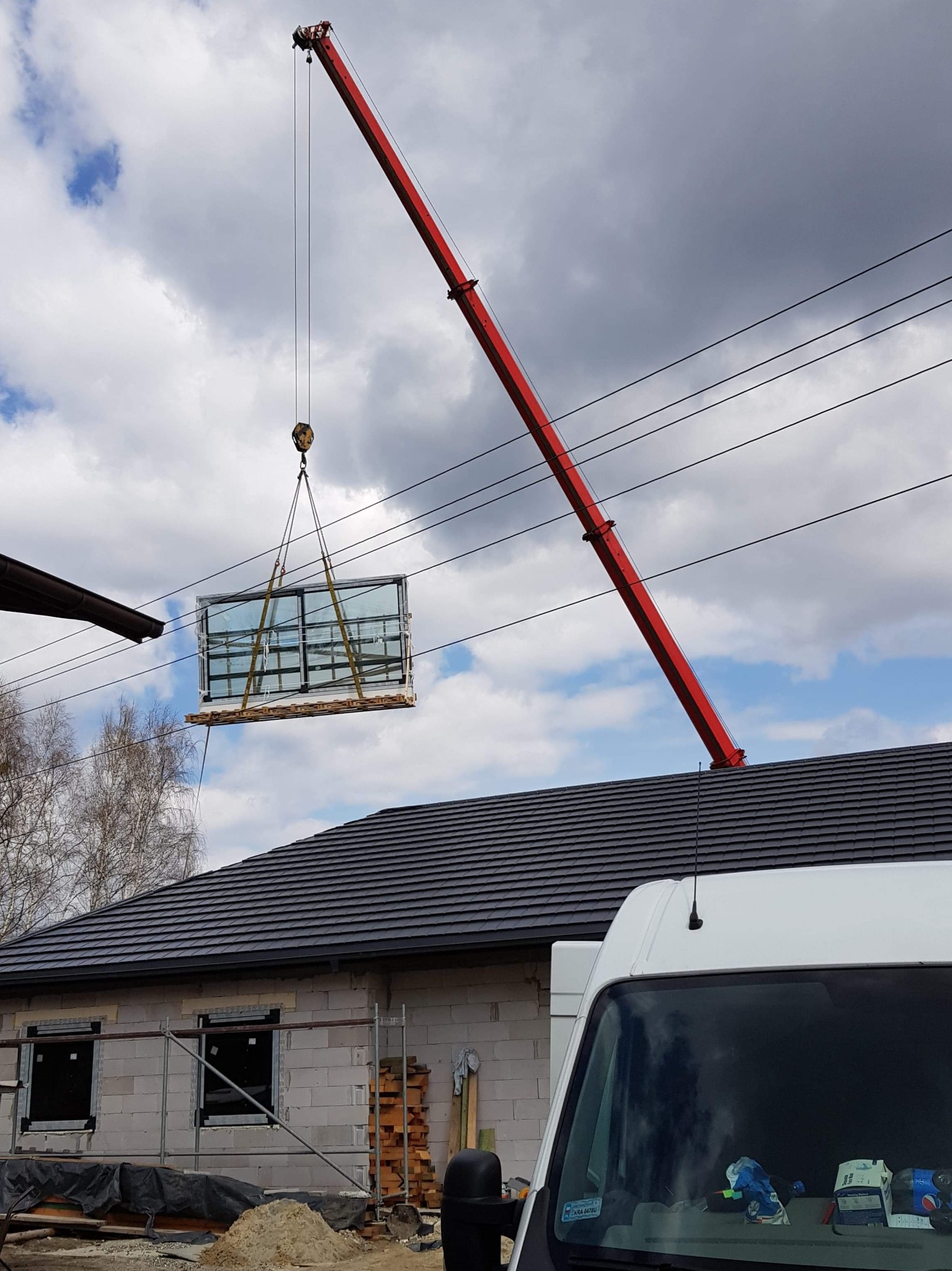 Czerwony dźwig w Gostyniu podnosi duże okno na palecie nad budynkiem w trakcie budowy, widoczny biały samochód dostawczy.