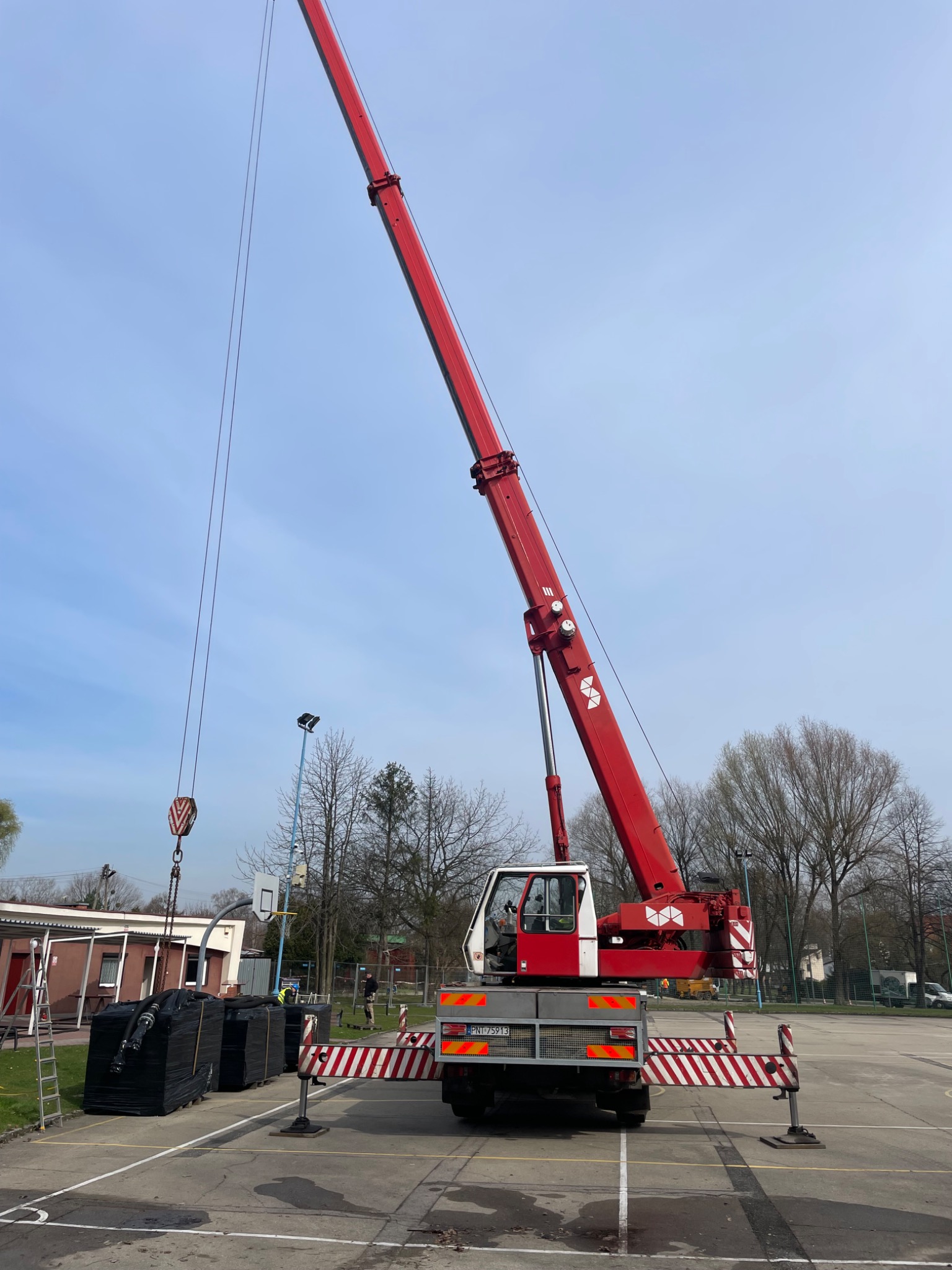 Czerwony dźwig samochodowy z wysuniętym ramieniem, przygotowany do podnoszenia czarnych pakunków na paletach na placu z widocznym boiskiem do koszykówki w tle, zabezpieczony barierkami ostrzegawczymi.