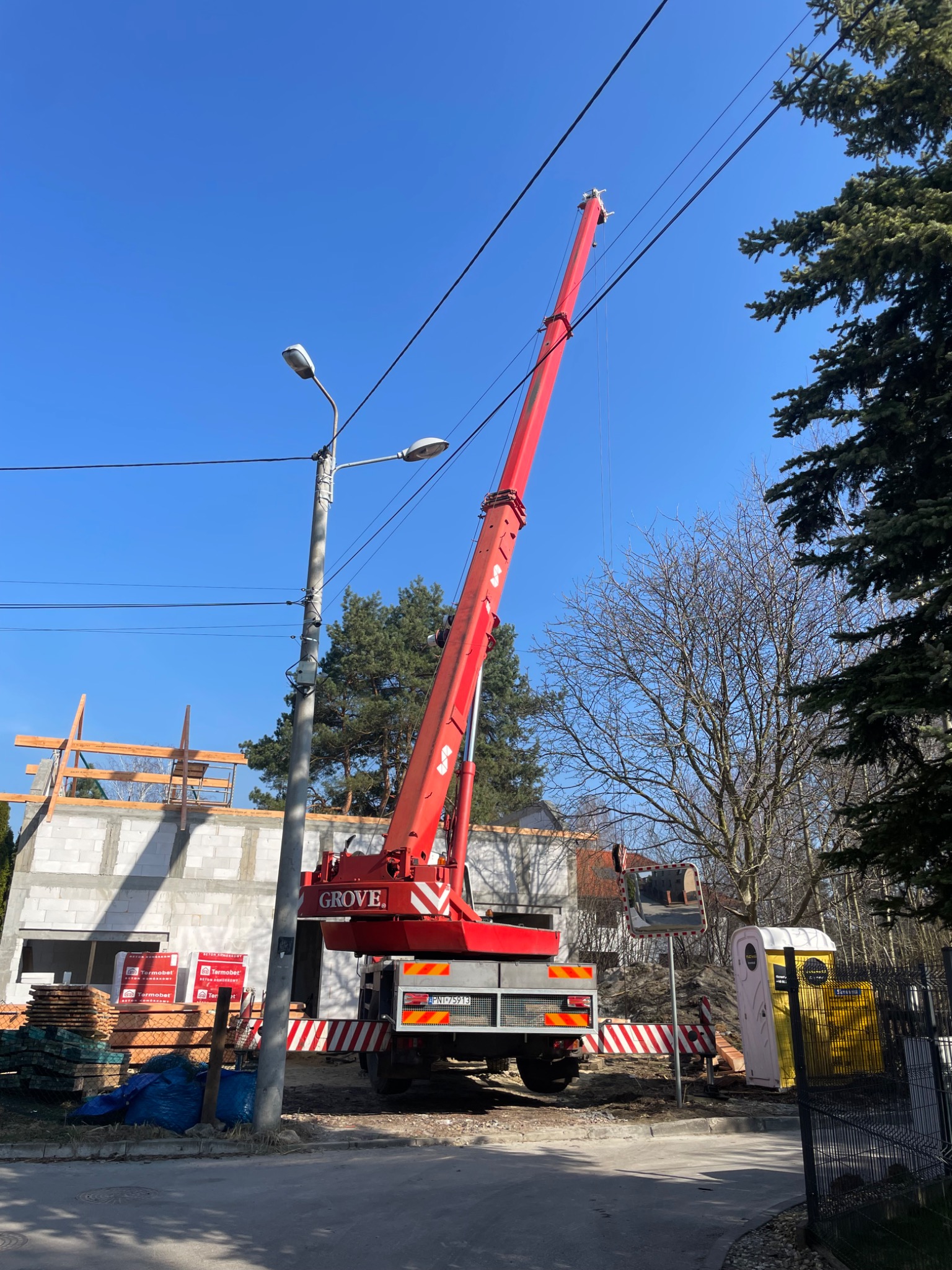 Czerwony dźwig Grove na tle budowy domu z bloczków, z widocznymi paletami materiałów budowlanych i przenośną toaletą.