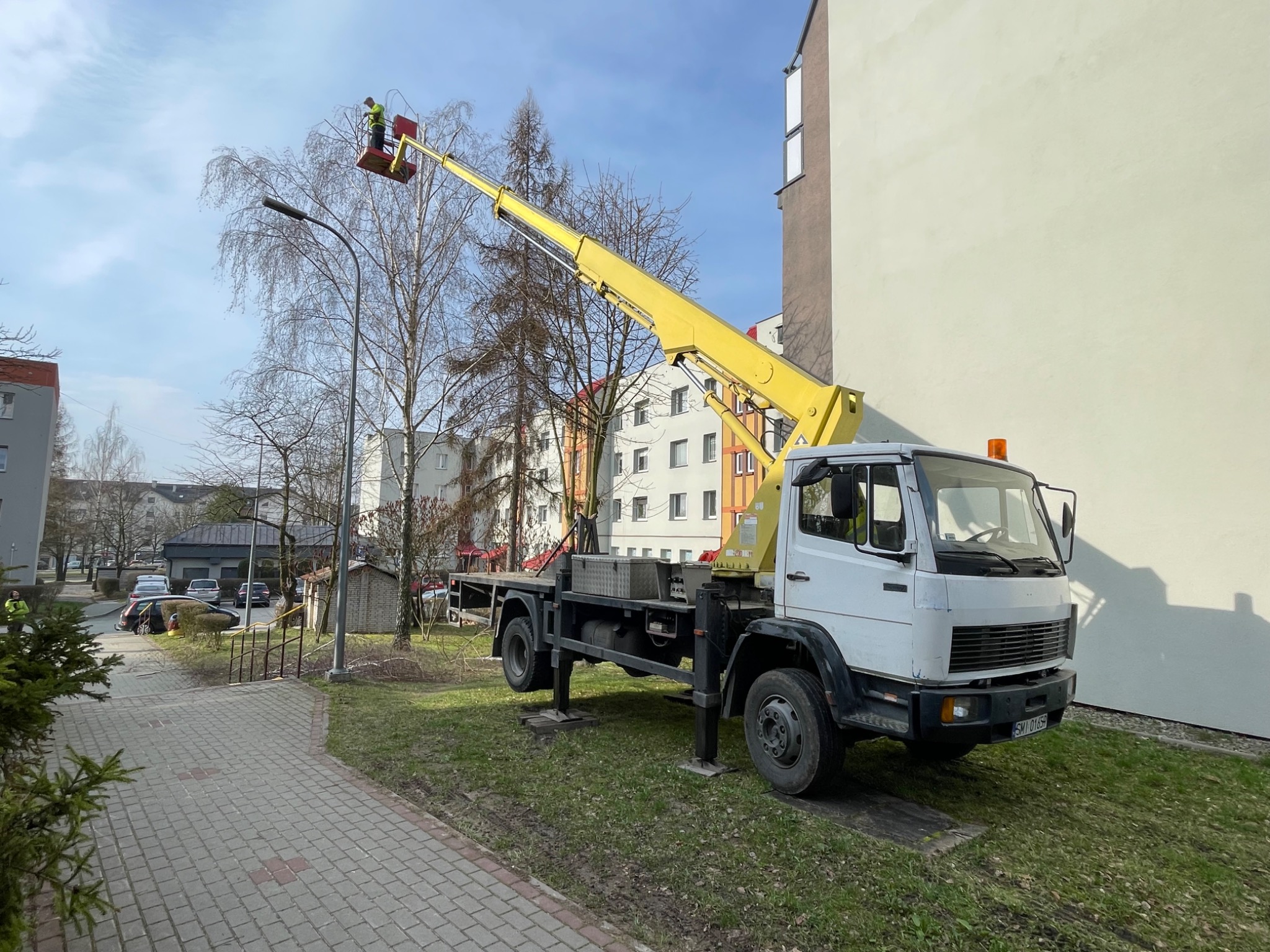 Żółty podnośnik koszowy na tle bloku mieszkalnego, pracownik w koszu przycina gałęzie drzewa, widoczne podpory stabilizujące pojazd.