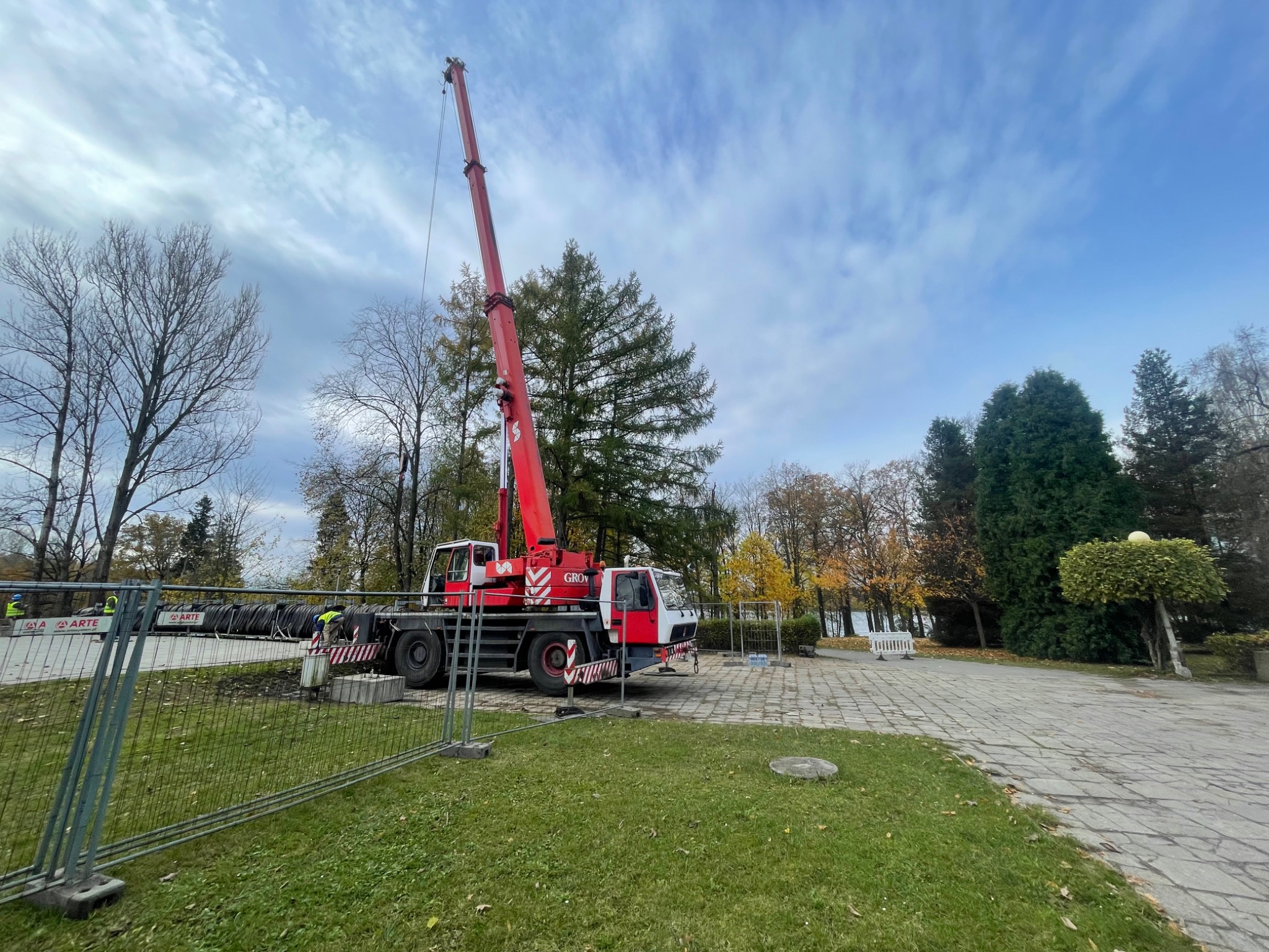 Czerwono-biały dźwig samochodowy marki GROV z wysuniętym ramieniem na tle pochmurnego nieba i drzew w parku, zabezpieczony ogrodzeniem tymczasowym.