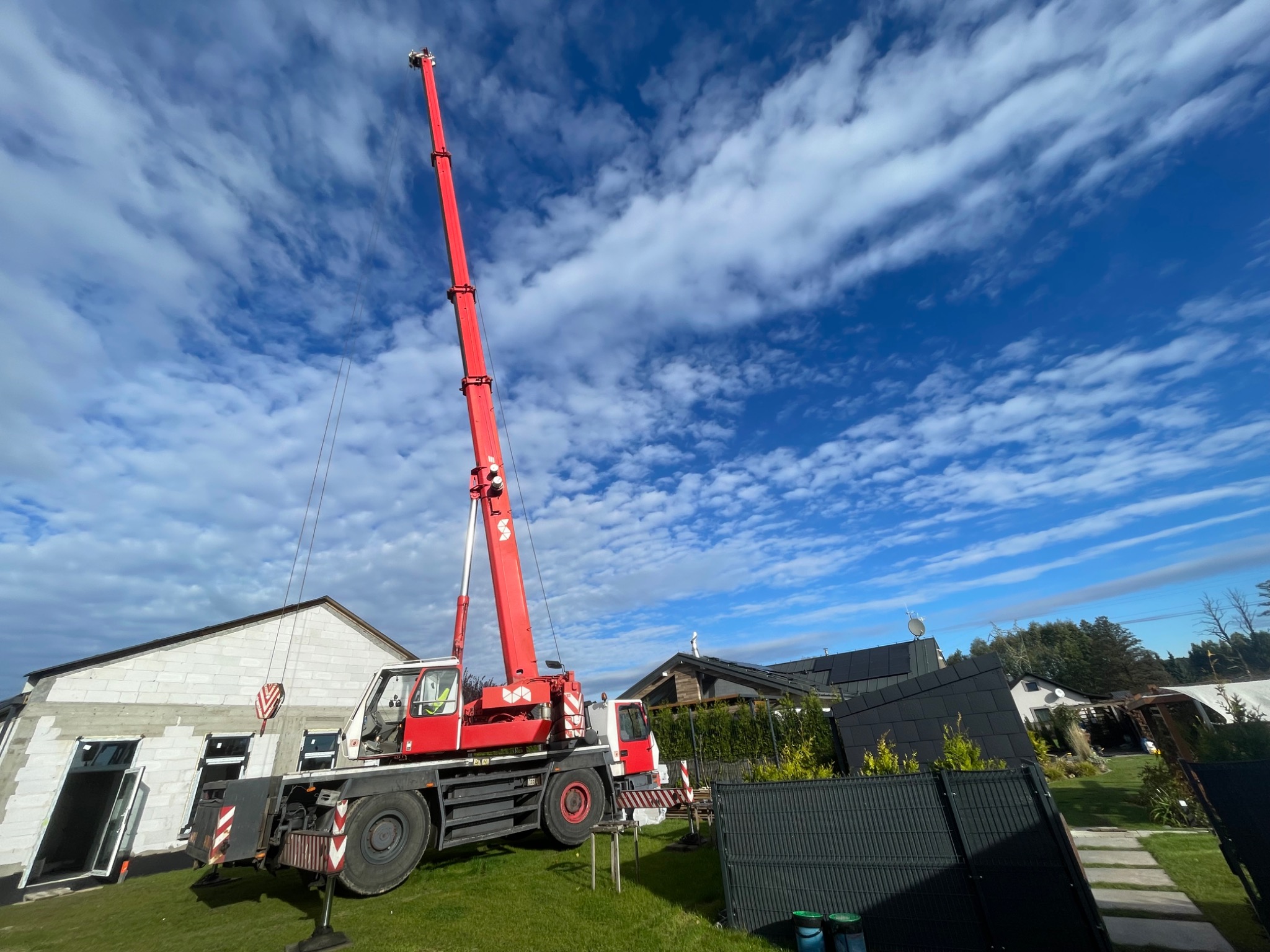 Czerwony dźwig samochodowy z wysuniętym ramieniem na tle błękitnego, pochmurnego nieba, pracujący przy budowie domu z białego bloczka