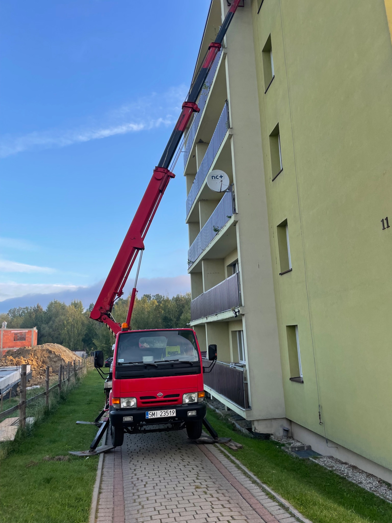 Czerwony dźwig samochodowy z wysięgnikiem sięgającym balkonu bloku mieszkalnego, widok z dołu na błękitne niebo